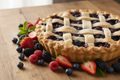 A delicious looking 9inch mixed berry pie with a flaky crust surrounded by mixed berries on a wooden table.