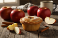 A delicious looking cupcake sized apple pie with a flaky crust surrounded by apples on a wooden table.