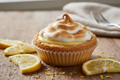 A delicious looking cupcake sized lemon meringue pie with a flaky crust surrounded by slices of lemon on a wooden table.